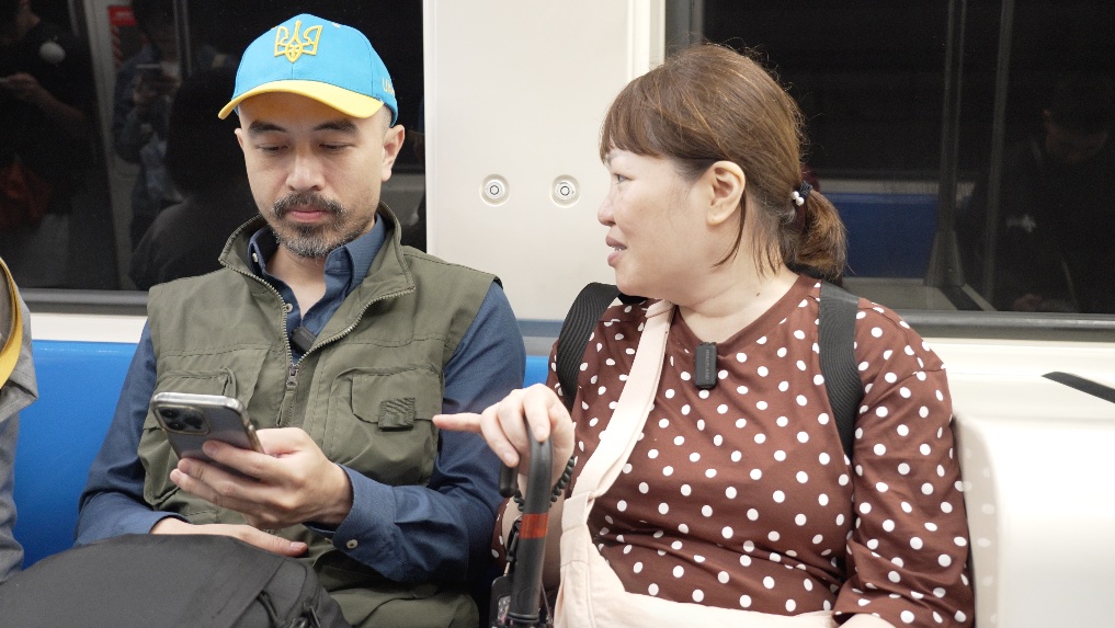 Long and Vi seated together on a metro train, looking at a phone.