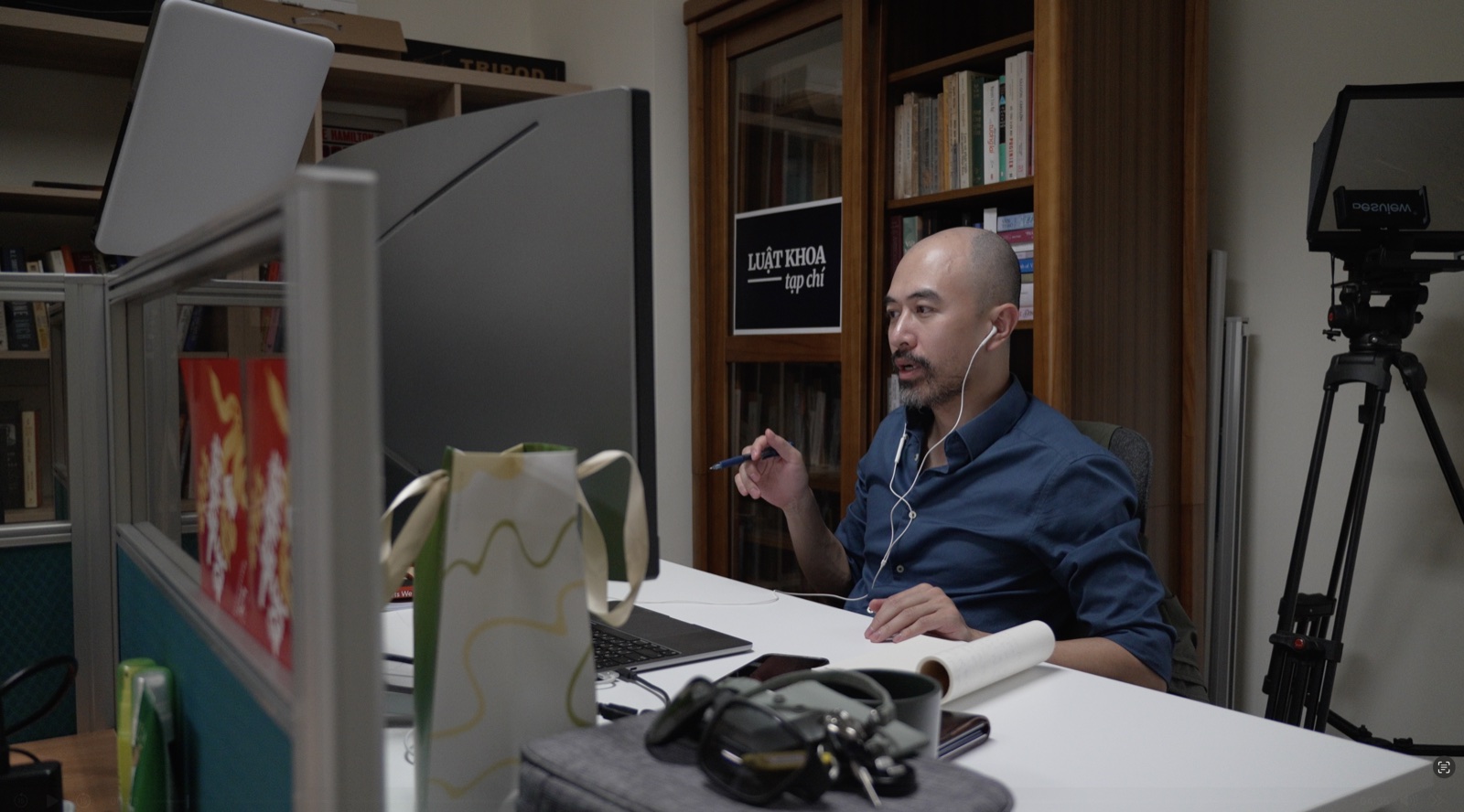 Trịnh Hữu Long at a desk in the Luật Khoa office, wearing earphones and holding a pen as he speaks on a video call, with a Luật Khoa tạp chí sign on the bookshelf behind him.